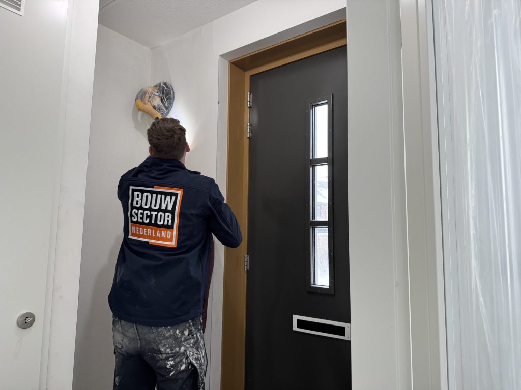 Stukadoor schuurt met een schuurmachine de bovenkant van een wand vlak onder het plafond van een hal