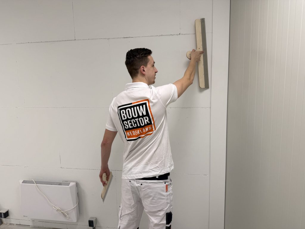 Stukadoor zet een lange spaan hoog tegen een wand naast een elektrische kachel