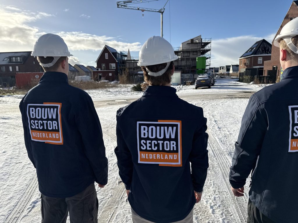 Drie medewerkers van Bouwsector Nederland in donkerblauwe jas en witte helm op een bouwterrein met nieuwe huizen